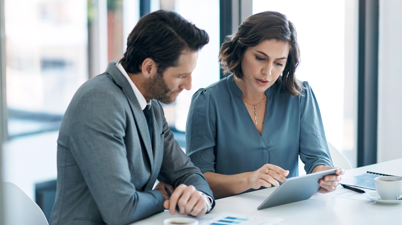 Due professionisti che discutono report finanziari in ufficio con documenti sul tavolo.